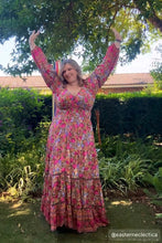 Woman in a floral boho dress standing outdoors with arms raised, surrounded by greenery.