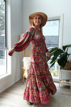 Woman in a boho floral dress and hat standing in a bright room with large windows.