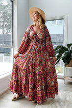 Woman in a colorful floral dress and wide-brimmed hat standing in a bright room with large windows.
