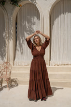 Woman in a brown dress standing in front of arched doorways with white curtains.