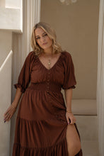 Woman wearing a brown boho dress standing indoors with neutral walls.