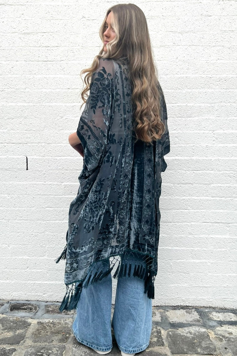 A woman wearing a long length boho poncho with floral design and tassels, in a steel grey color, paired with denim and boots.
