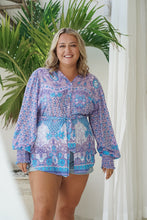 Woman wearing a colorful boho patterned outfit standing in a bright room with plants.