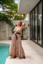 Woman in a long, patterned dress standing by a pool with modern architecture and greenery in the background.