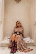 Woman in a floral dress and cowboy boots sitting on a bench in a classical architecture setting.