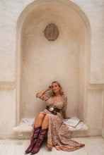 Woman in a floral dress and cowboy boots sitting in an arched stone alcove.