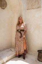 A person wearing a caramel floral maxi dress with balloon sleeves and a low v-neck, styled with a brown belt and boots, standing in a bohemian-themed setting.