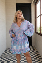 Woman wearing a blue boho patterned dress standing in a room with checkered floor and window.