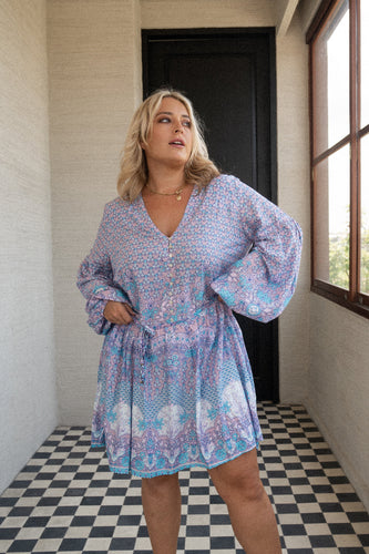 Woman wearing a blue boho patterned dress standing in a room with checkered floor and window.