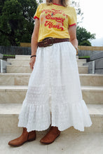 Person wearing a yellow shirt with red text, white skirt, and brown boots on stone steps.