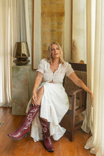 Woman in a white boho skirt and red cowboy boots sitting on a wooden chair in a room with curtains and a lamp.