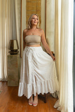 Woman wearing a beige top and white boho skirt standing in a room with curtains and a lamp.
