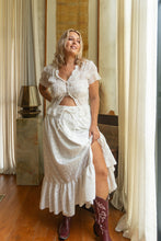 Woman in a white dress with a slit standing in a room with curtains and a lamp.