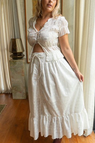 Woman wearing a white boho dress with ruffled sleeves and a tied waist in a room with curtains and a lamp.