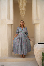 Woman in a boho floral dress standing in a decorative interior setting with a chandelier and large vase.