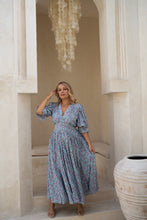 Woman in a boho floral dress standing in a sunlit room with architectural columns.