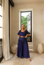 Woman in a boho blue outfit standing in a room with large windows and decorative vases.