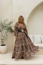 Woman in a boho leopard print dress standing in a stylish interior setting with a plant and sofa.