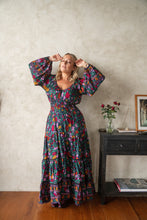 Woman wearing a colorful boho floral dress standing in a room with a table and vase.