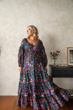 Woman wearing a colorful floral boho dress standing in a room with a neutral wall and wooden floor.