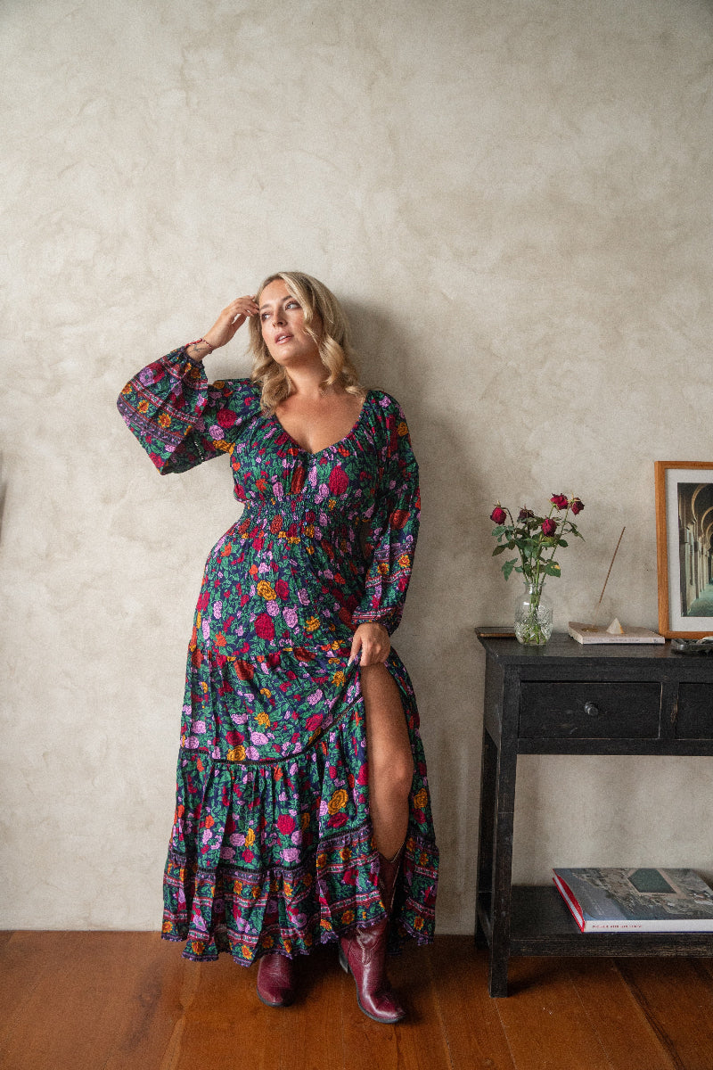 A woman standing in an indoor setting wearing a long, boho floral print maxi dress with long balloon sleeves and a low v-neck.