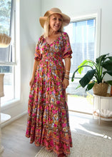Woman in a boho floral dress and hat standing in a bright room with large windows.
