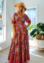 Woman in a boho floral dress and wide-brimmed hat standing in a bright room with large windows.