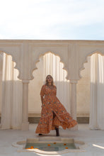 Woman in an orange boho floral dress standing in a sunlit room with arched windows and a fountain.