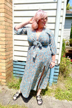 A woman wearing a boho floral print front tie dress in blue and beige colors, with an above-knee split and elbow-length sleeves, standing against a brick wall and wooden fence background.