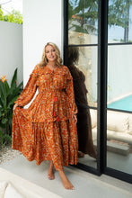 A woman standing by a glass door wearing a boho rust-colored paisley print maxi dress with a voluminous skirt and balloon sleeves.