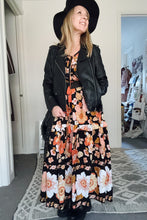 Woman wearing a floral boho dress with a black jacket and hat indoors.