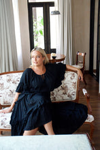 Woman in a black dress sitting on a floral-patterned chair in a room with large windows.