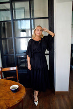 Woman in a black dress standing in a room with a wooden table and chair.