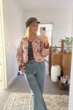 Person taking a mirror selfie wearing a boho patterned jacket, jeans, and a cap in a room.