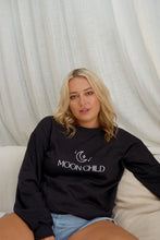 A person wearing a black boho sweatshirt with the text 'MOON CHILD' on it, sitting on a couch.