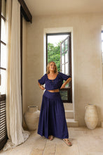 Woman in a boho blue outfit standing in a room with large windows and beige walls.