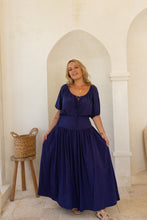 Woman in a boho blue dress standing in a sunlit room with arched doorways.