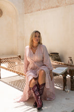 Woman in a pink boho dress and cowboy boots sitting on a wooden bench in a sunlit room.