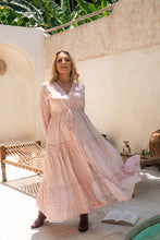 Woman in a long, light pink boho dress standing in an outdoor setting with a white wall and wooden furniture.