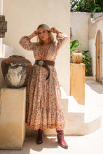 Woman in a patterned boho dress with a belt and boots standing in a sunlit outdoor setting.