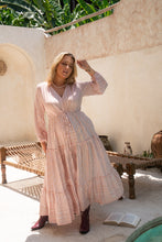 A woman standing outdoors wearing a pink boho maxidress with beige toned shells and a border print, paired with boots and a headscarf.