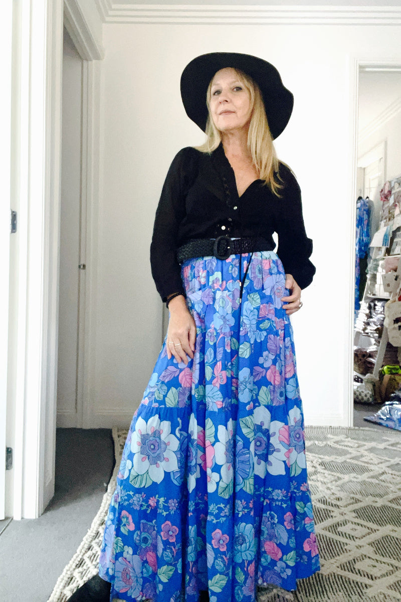 Woman wearing a black top and blue floral boho skirt with a black hat indoors.