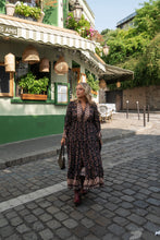 Woman in a floral boho dress walking on a cobbled street in front of a green building with plants.