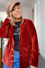 A person wearing a boho heritage brown velvet blazer with a collar, pointed lapels, and front pockets. The blazer is short-length and the person is also wearing a black top and blue jeans.