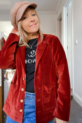 A person wearing a boho heritage brown velvet blazer with a collar, pointed lapels, and front pockets. The blazer is short-length and the person is also wearing a black top and blue jeans.