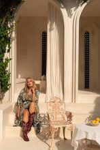 Woman in a boho floral dress and red boots sitting on steps in an elegant outdoor setting with columns and a chair.