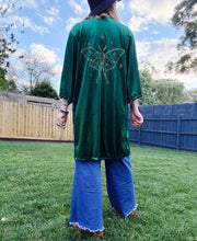 A woman standing outdoors on a grassy lawn, viewed from behind, wearing a dark green velvet tunic with a large gold butterfly design on the back, blue wide-leg jeans, and a dark hat.
