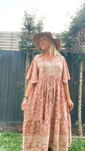 A woman wearing a floral boho dress and a hat, standing outside the garden looking in the side.