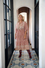 Woman in a pink floral dress standing in a doorway with a patterned floor.