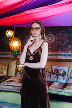 Woman in a dark brown boho dress with floral embroidery in a colorful room with hanging lights and patterned cushions.